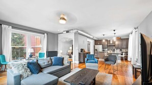 A modern living room with a grey sofa, blue armchairs, and a wooden coffee table. A kitchen with dark cabinets and pendant lights is visible in the background. - 8 min to Stadiums & Dwntn Cincy, 1BR in Ludlow KY (Ludlow)