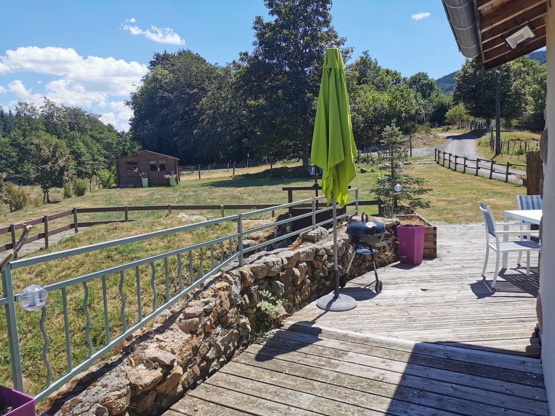A wooden deck with outdoor seating, a barbecue grill, and a view of a rural landscape with trees and a small building.