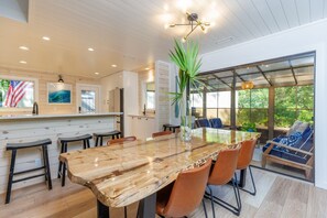 A modern kitchen with a large wooden table and bar stools, a hanging light fixture, and large windows offering a view of the outdoors. - 6 Pine Court: Modern Sea Pines Home, Community Pool, Tennis, Free Bikes (Hilton Head Island)