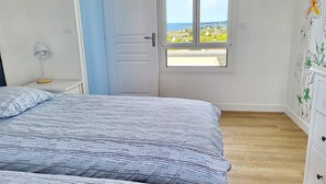 Ein Schlafzimmer mit Bett, Nachttisch, Lampe, Tür und Fenster mit Blick auf das Meer und Häuser.