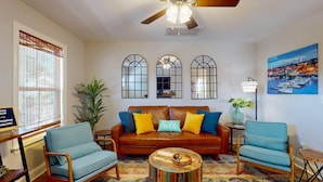 A living room with a brown leather sofa, two blue armchairs, a wooden coffee table, and a colorful rug. There are three arched mirrors on the wall, a ceiling fan, and a painting of a harbor. - Mins to Downtown, Live Oak Concerts, Convention Center & Beach! Pet Friendly!! (Wilmington)