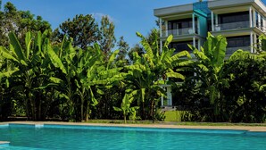 A swimming pool surrounded by lush banana trees and a modern building with balconies. - Shose Farmhouse Chalets & Campsite (Moshi)