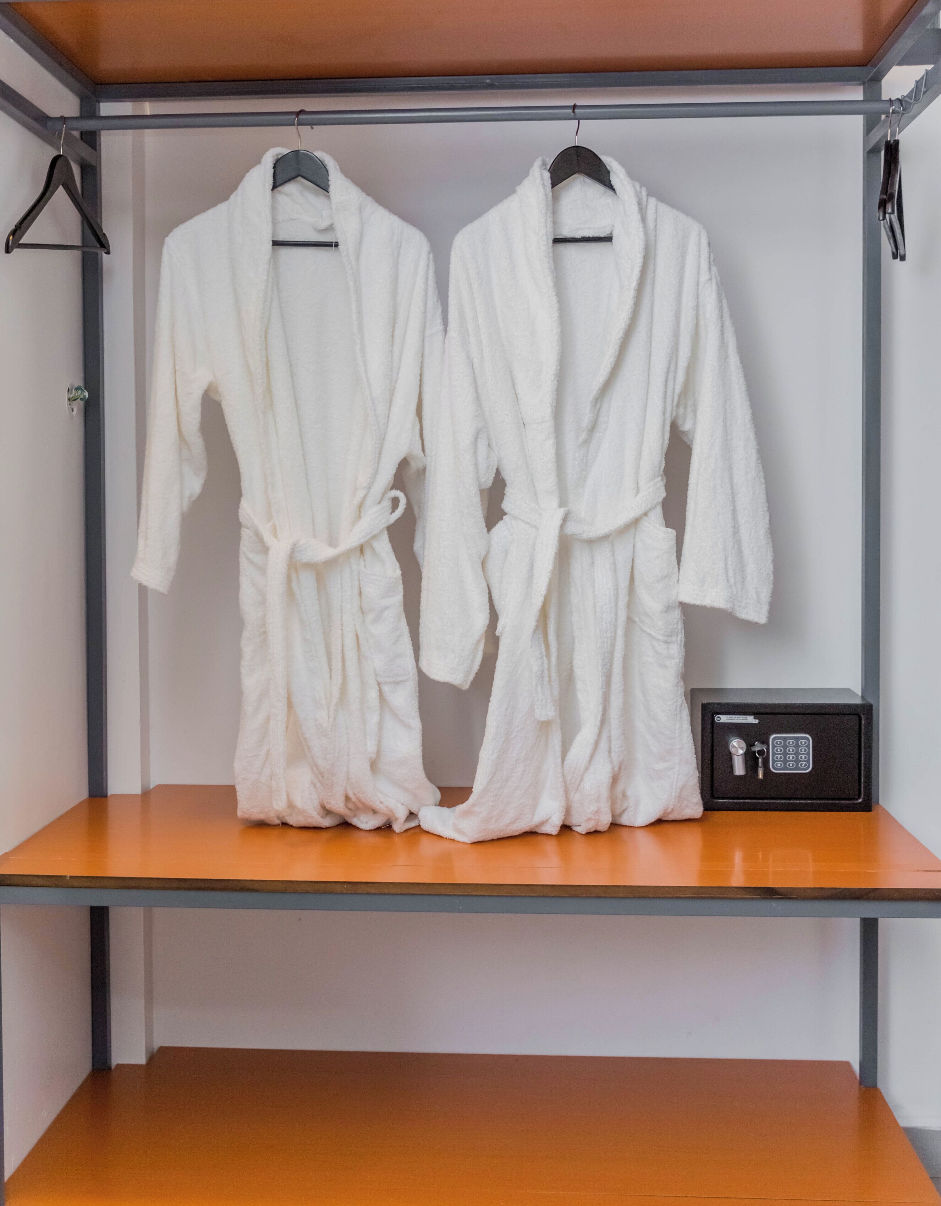 A storage unit with two white robes hanging on hangers and a safe on the shelf.