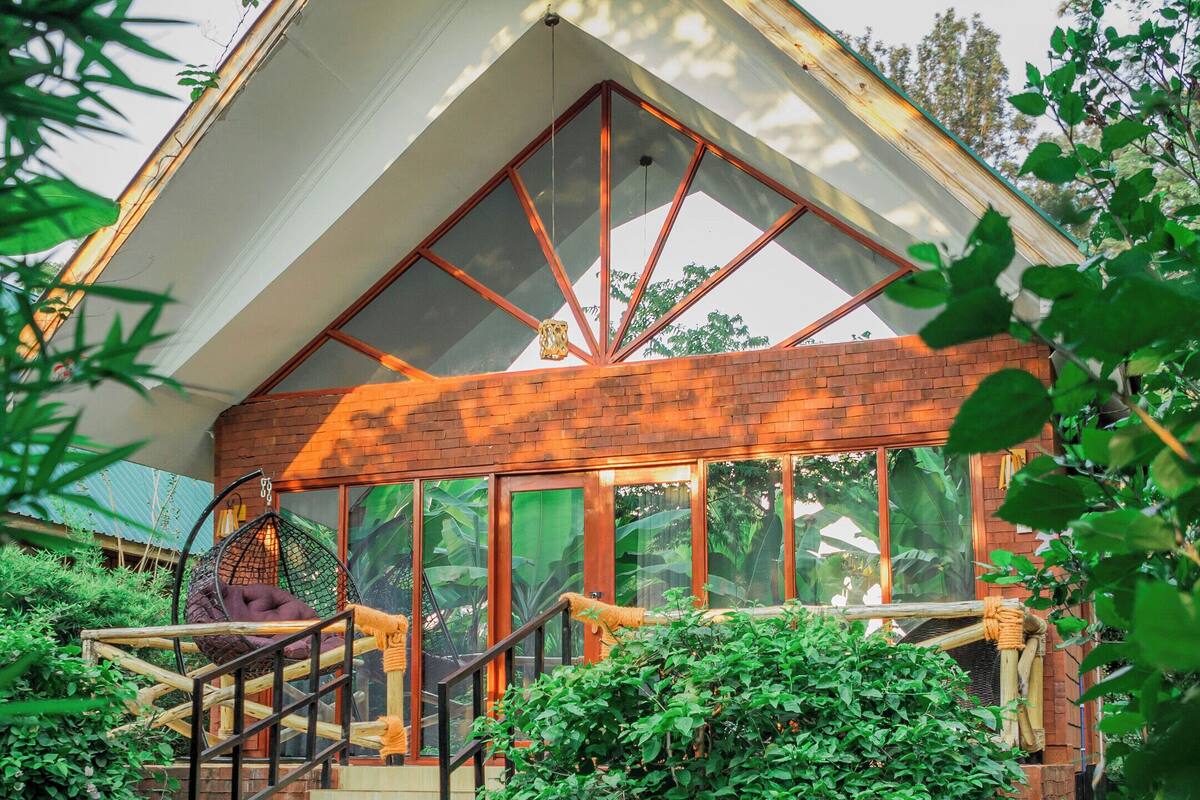 A modern building with a sloped roof, a wooden staircase leading to a covered porch, and a surrounding lush garden.
