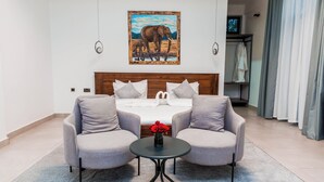 A hotel room with a bed, two armchairs, a small table with a vase of flowers, and a painting of elephants on the wall. - Shose Farmhouse Chalets & Campsite (Moshi)