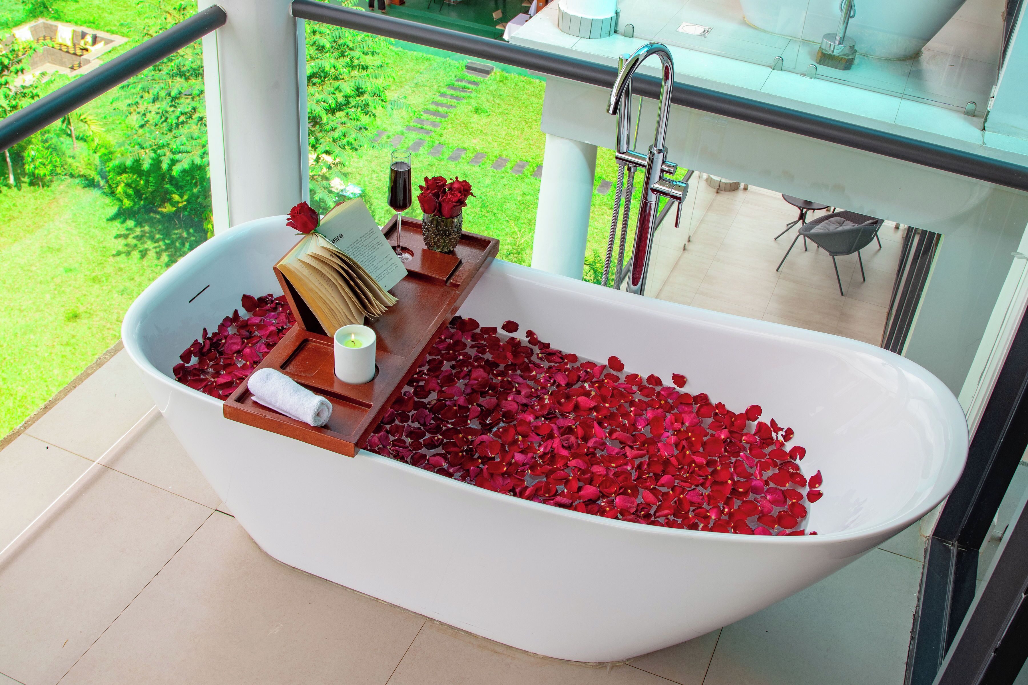 A modern bathroom with a freestanding bathtub filled with rose petals, a glass of wine, and a book on a wooden tray.