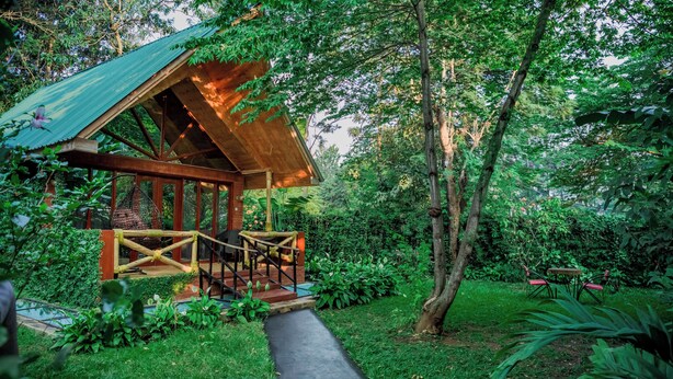 A wooden cabin with a porch, surrounded by lush greenery and trees. - Shose Farmhouse Chalets & Campsite (Moshi)