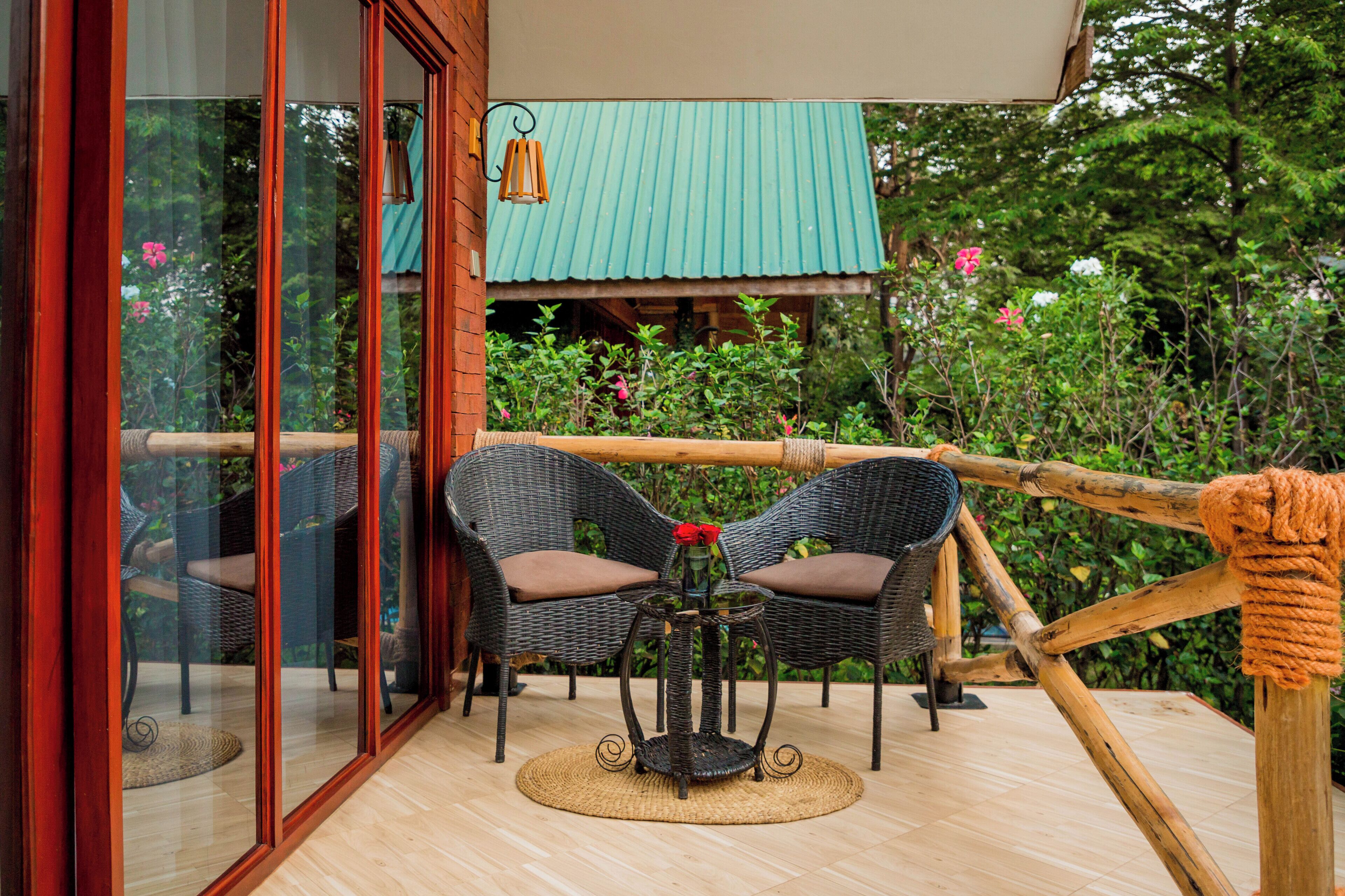 A glass-enclosed balcony with wicker furniture, a small table with a vase, and a view of a green-roofed building.
