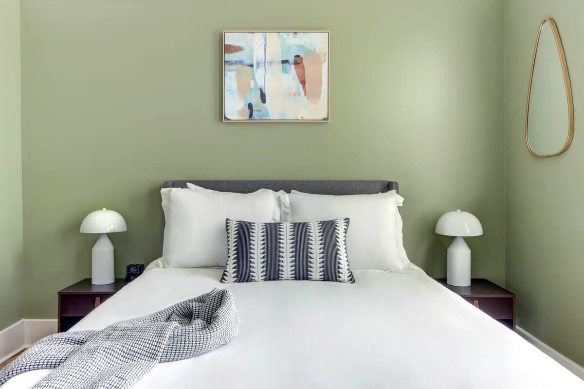 Una cama bien hecha con sábanas blancas y almohadas estampadas, dos lámparas de noche y un cuadro abstracto enmarcado en la pared.