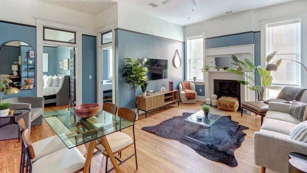 A spacious living room with a glass coffee table, a fireplace, and a television mounted on the wall. - Tidewater Blue @ The Huntingdon Lofts Collection (Savannah)