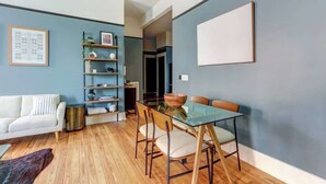 A modern living room with a glass dining table, wooden flooring, a white sofa, and a bookshelf. - Tidewater Blue @ The Huntingdon Lofts Collection (Savannah)