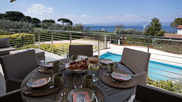 An outdoor dining area with a view of the sea, equipped with tables, chairs, and a pool.