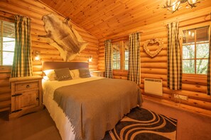 Ein Schlafzimmer in einer Blockhütte mit einem Bett, Nachttischen, einem Fenster mit Vorhängen, einer Wandheizung und einem Hirschfell an der Wand.