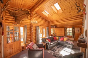 A log cabin interior with a leather sectional sofa, wooden walls, and a mounted deer head.