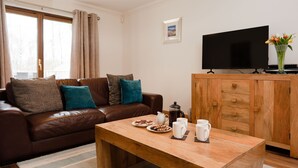 A cozy living room with a brown leather sofa, a wooden coffee table with snacks and mugs, a TV on a wooden stand, and a cabinet with drawers.