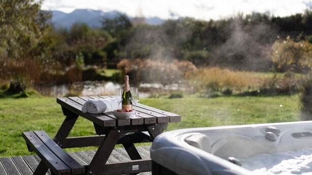 Una mesa de picnic con una botella y vasos, un cuenco de fresas y un jacuzzi en un entorno natural con montañas al fondo.