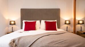 A neatly made bed with a grey headboard, two red pillows, and a beige throw blanket. There are two bedside tables with lamps on either side of the bed. - sleeps 6 in 1 double bedroom, 1 twin (or superking), 1 full size bunk bed. (Aviemore)