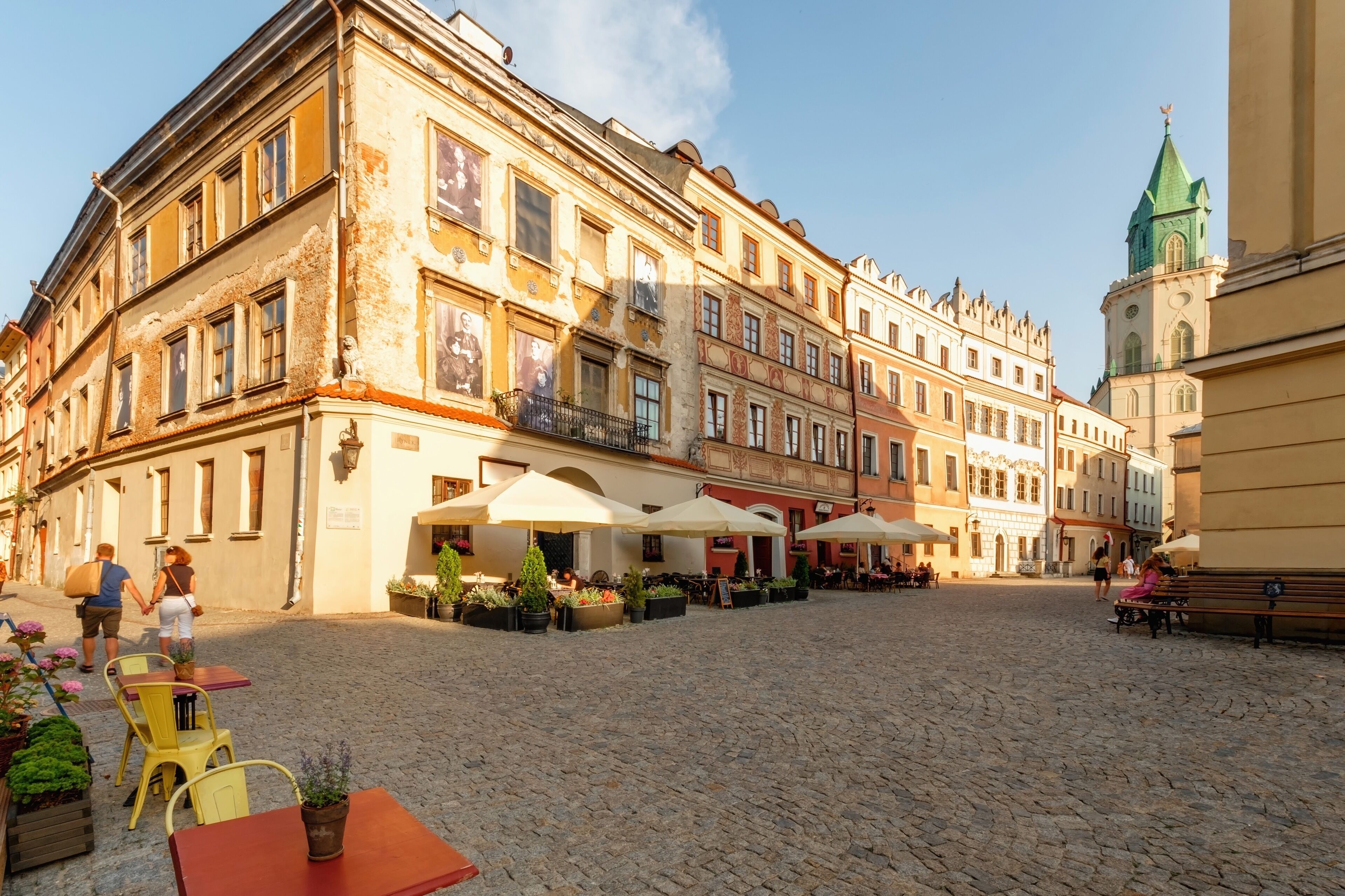An outdoor café area with tables and chairs, surrounded by historic buildings with balconies and a clock tower.