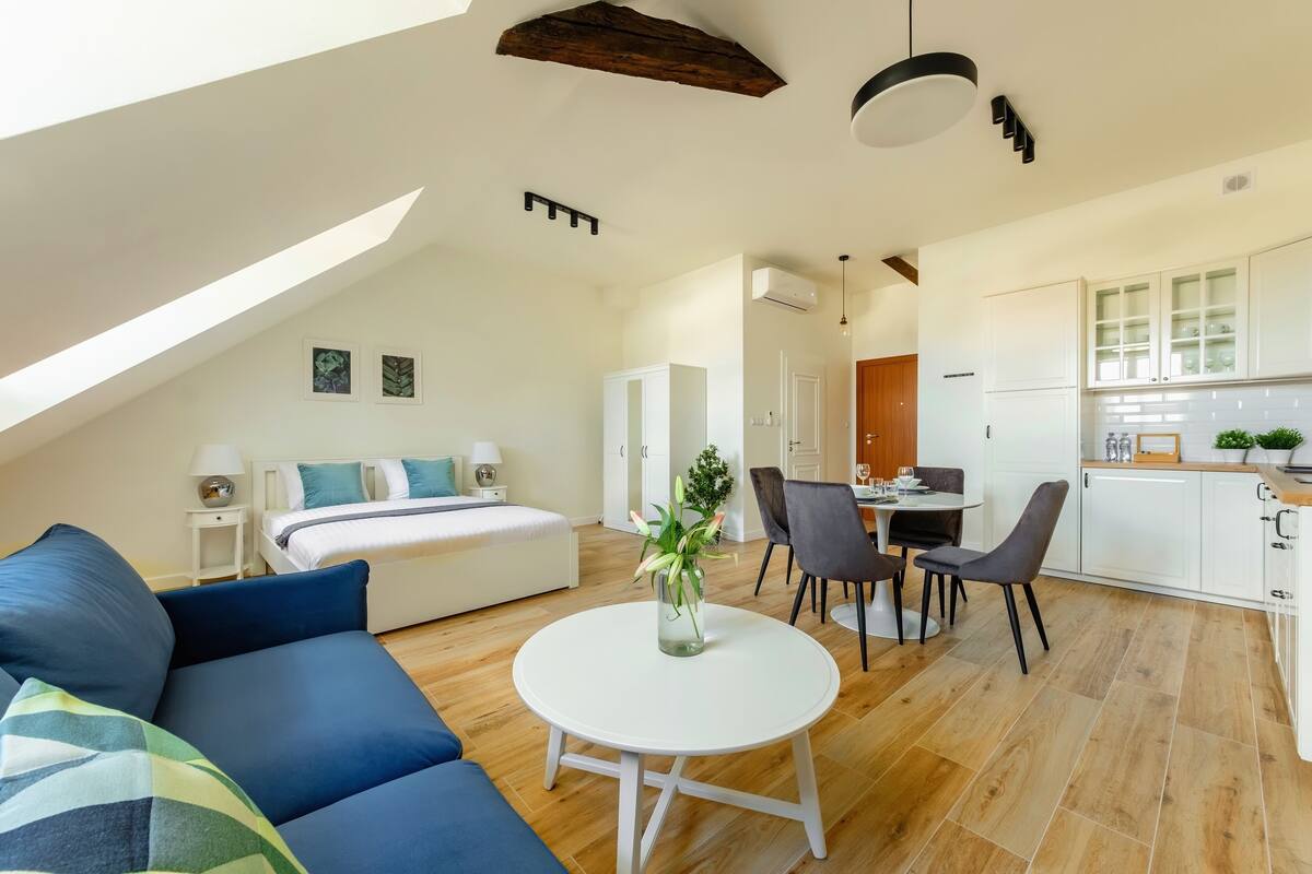 A modern, well-lit room with a bed, a blue sofa, a dining table with chairs, and a kitchen area.