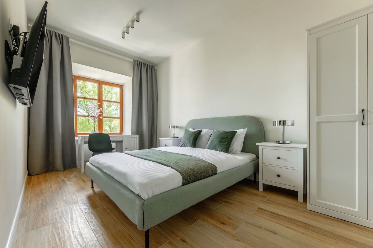 A bedroom with a bed, two bedside tables, a desk, a chair, a wardrobe, and a TV mounted on the wall.