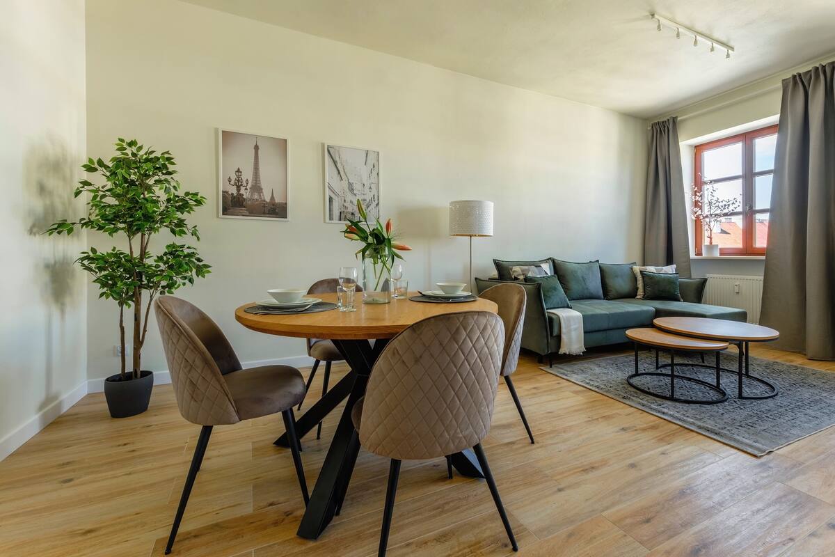 A modern living room with a dining area, featuring a round wooden table, cushioned chairs, a sofa, and a potted plant.