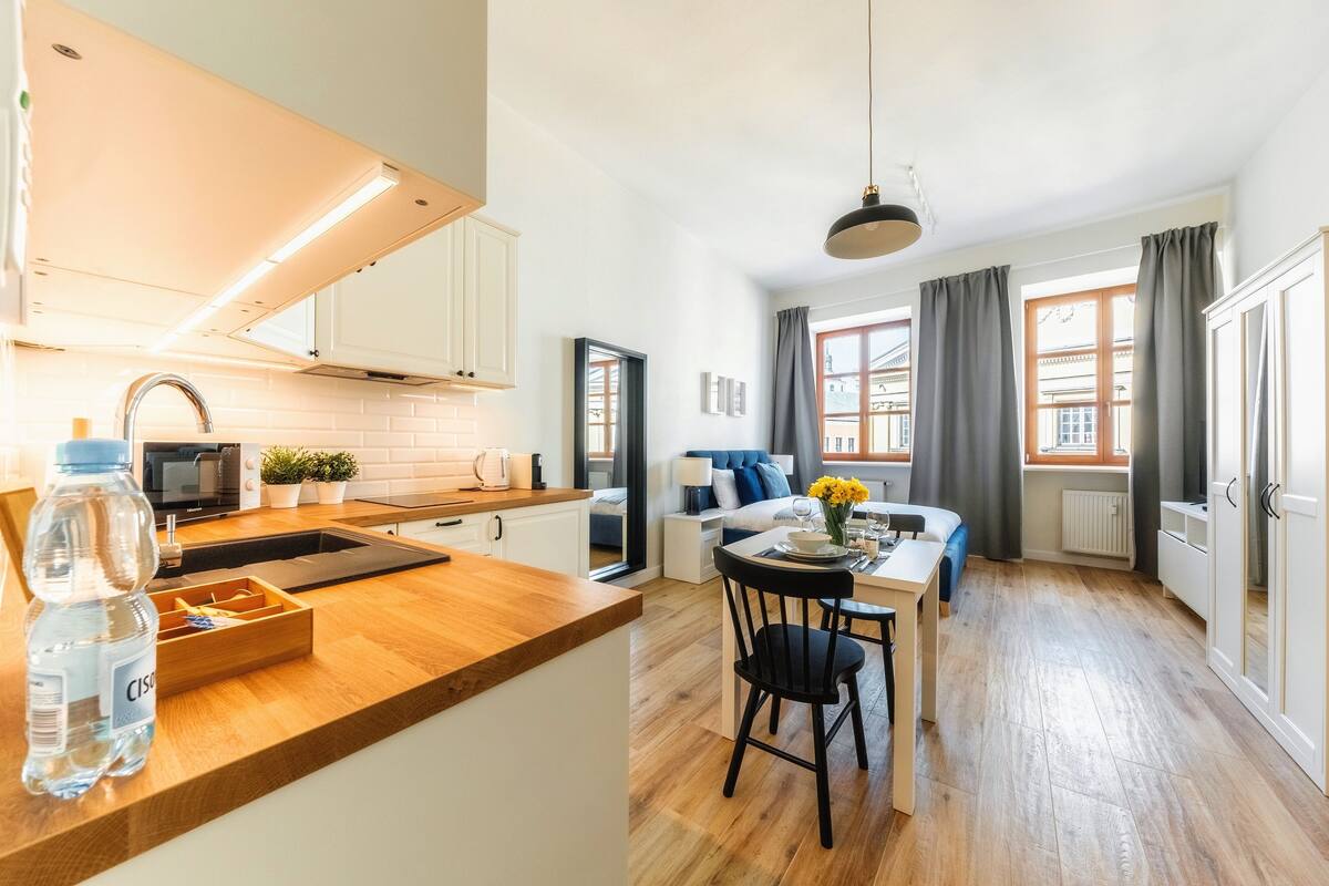A modern kitchen with a wooden countertop, a sink, and a water bottle. There is a dining area with a table and chairs, and a sofa in the background.