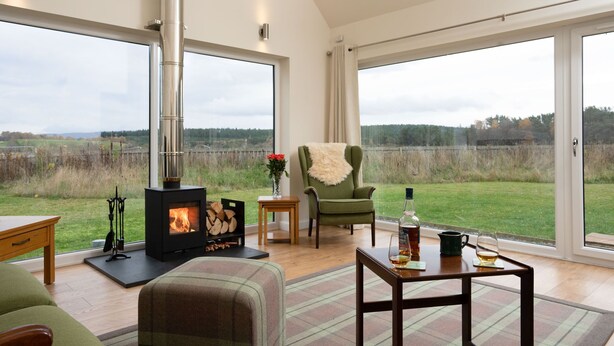 A spacious room with a fireplace, a green armchair, a table with drinks, and a view of a grassy field through large windows. - Sleeps 6 in 2 doubles and 1 twin room (Dulnain Bridge)