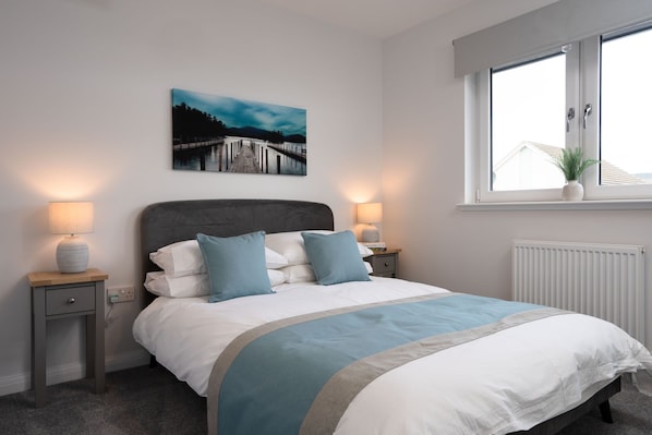 A bedroom with a bed, two bedside tables, a nightstand, a window with a view, a radiator, and a painting on the wall.