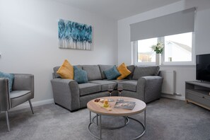 A modern living room with a grey sectional sofa, a round coffee table with snacks and wine glasses, a television, and a large window with a view of houses.