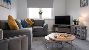 A modern living room with a grey sofa, a round coffee table with food and drinks, a flat-screen TV, and a window with a view of greenery.