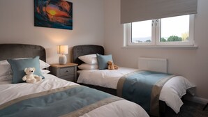 A bedroom with two beds, a nightstand with a lamp, a teddy bear, a painting, and a window with a view.