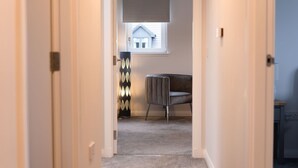 A narrow hallway with a carpeted floor, a window with blinds, a chair, and a decorative wall panel.