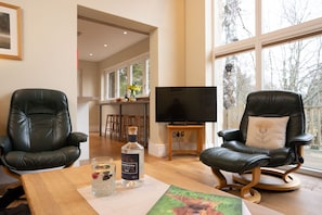 A living room with two leather chairs, a wooden coffee table with a glass of gin and a coaster, a television on a stand, and a large window with a view of trees. - Sleeps 12 in 6 double bedrooms all en-suite (Aviemore)