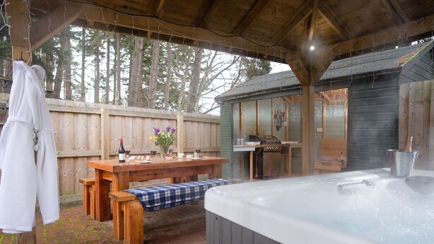 Ein überdachter Außenbereich mit Whirlpool, Holztisch und Grill.