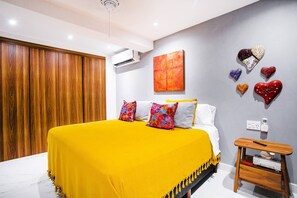 A bedroom with a bed, pillows, a bedside table, a wall-mounted air conditioner, a painting, and decorative heart-shaped objects.