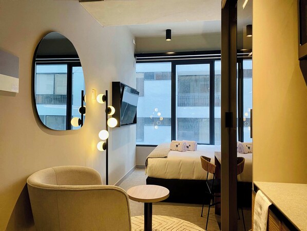 Une chambre d’hôtel moderne avec un grand lit, un grand miroir rond doté de lumières, une télévision et une vue sur la ville.