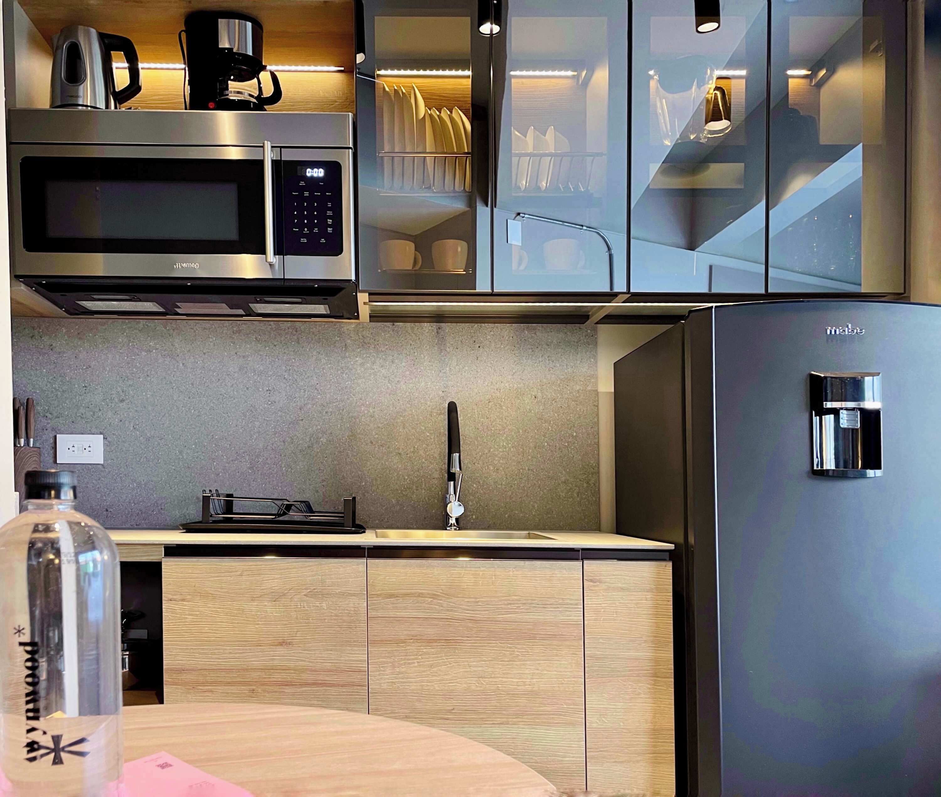 Une cuisine moderne équipée d’un four à micro-ondes en acier inoxydable, d’une machine à café, d’un distributeur d’eau et de meubles de rangement en bois.