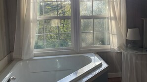 A bathroom with a large bathtub, a window with curtains, and a small table with a lamp.