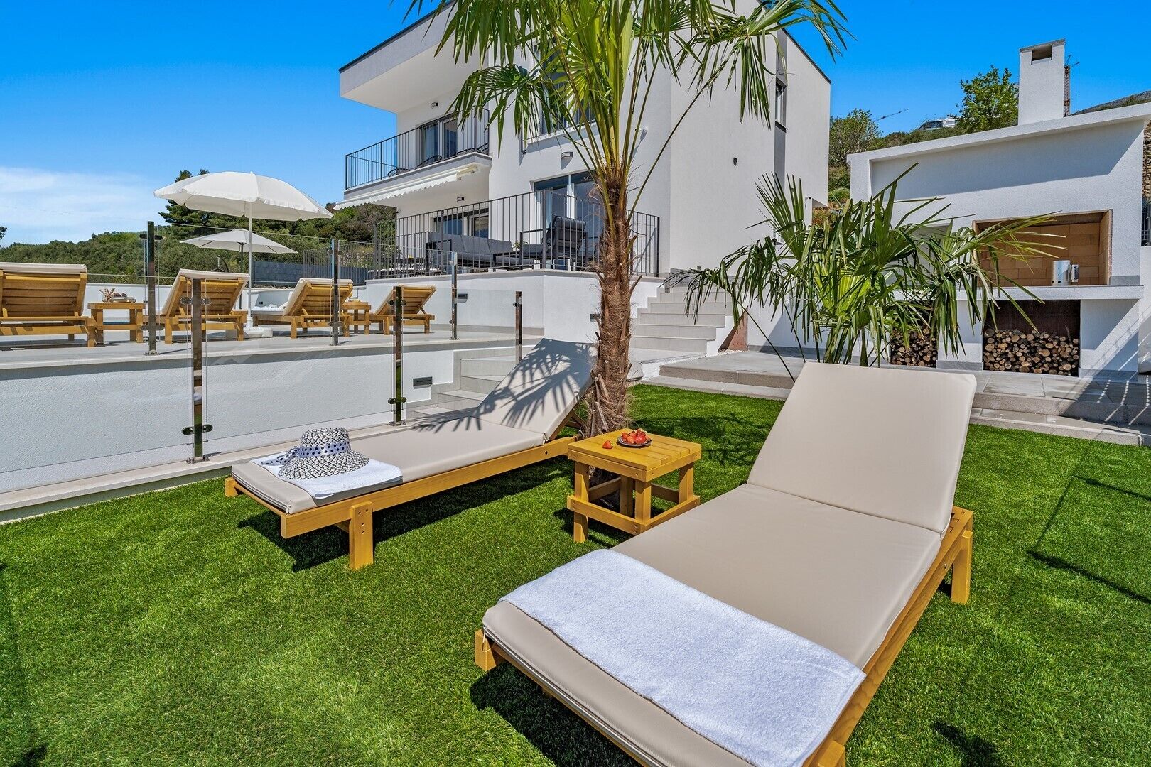 A modern outdoor lounge area with sun loungers, a small table with a bowl of fruit, and a staircase leading to a second-floor balcony.