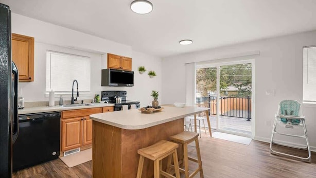 A modern kitchen with wooden cabinets, a central island with stools, a built-in oven, and a large window overlooking a balcony. - Family Friendly Haven I  Renovated & Spacious! (Spokane Valley)