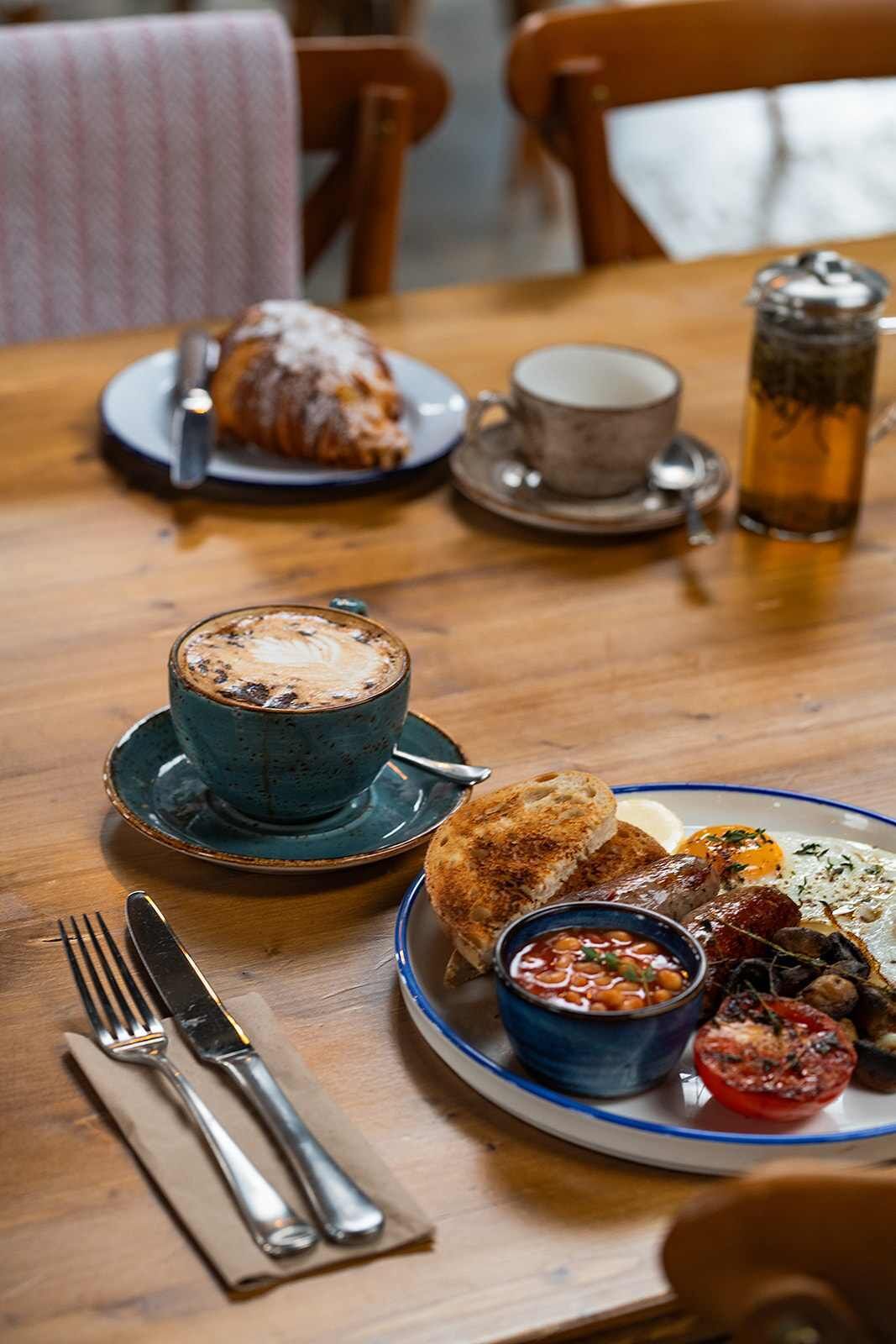 Ein Tisch gedeckt mit einem Croissant, einer Tasse Kaffee, einem Glas Eistee, einem Teller mit gebackenem Tomaten und einer Schüssel Bohnen.