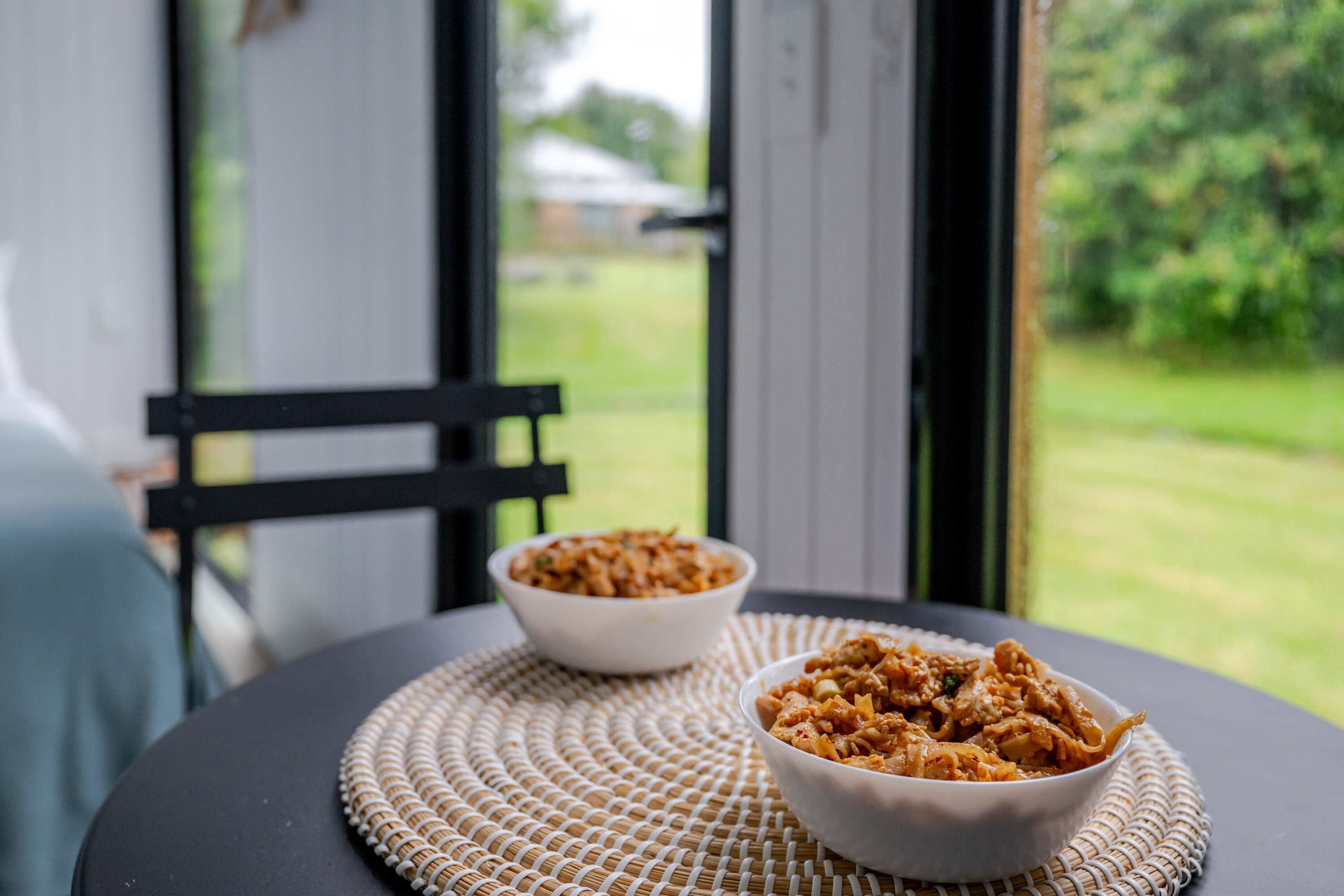 Two bowls of food on a table with a woven placemat, a chair, and a view of a green landscape through a window.