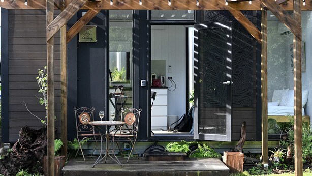 Une pergola en bois avec une table et des chaises, un petit espace jardin et une porte donnant sur une pièce à l’intérieur blanc.