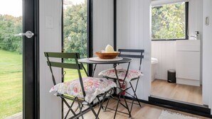 Une pièce donnant sur un paysage verdoyant, une petite table ronde avec un bol et une bougie, deux chaises avec des coussins à motifs floraux et un lavabo blanc.