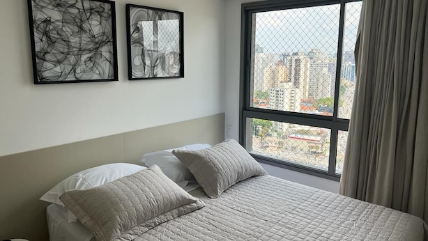 A bedroom with a large bed, two pillows, a nightstand, and a window with a view of buildings. - Flat Itaim SP (São Paulo)