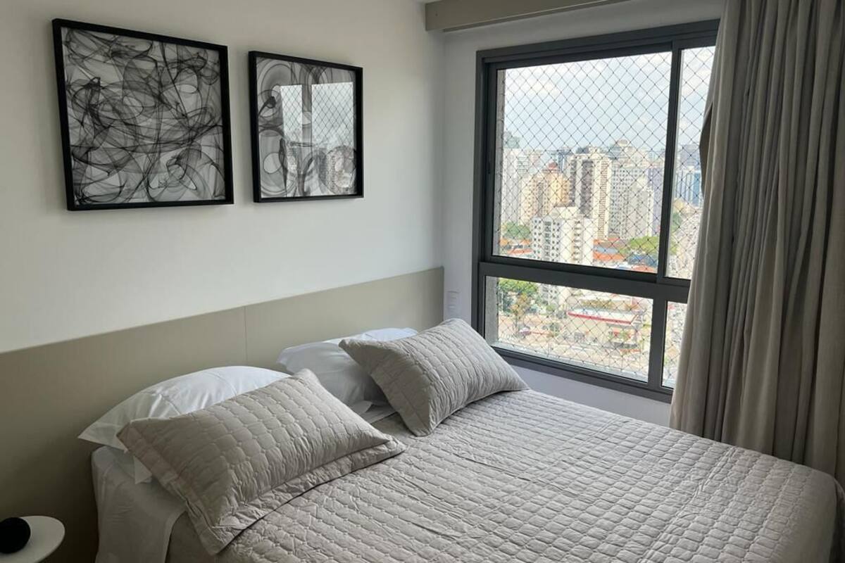 A bedroom with a large bed, two pillows, a nightstand, and a window with a view of buildings.