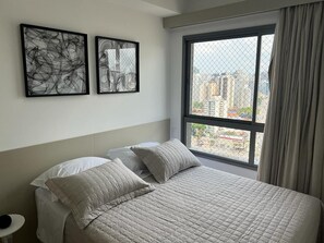 A bedroom with a large bed, two pillows, a nightstand, and a window with a view of buildings. - Flat Itaim SP (São Paulo)