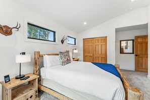 Een slaapkamer met een groot bed, houten hoofdeinde, nachtkastje met een lamp, een lijst met een afbeelding en een raam met uitzicht op bomen.
