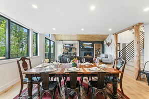 Een eetgedeelte met een grote houten tafel voor zes personen, omringd door donkere stoelen. Achtergrond is een open haard zichtbaar, en rechts een trap.