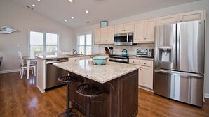 A modern kitchen with a central island, stainless steel appliances, and a dining area with a white table and chairs. - Surf N Dolphin, Oceanview House in Emerald Isle, NC! (Emerald Isle)